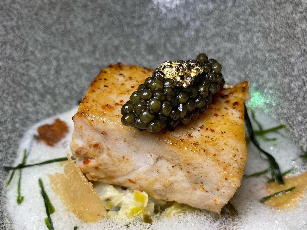 A large piece of hamachi, topped with caviar and goldleaf, all sitting atop a white foam.