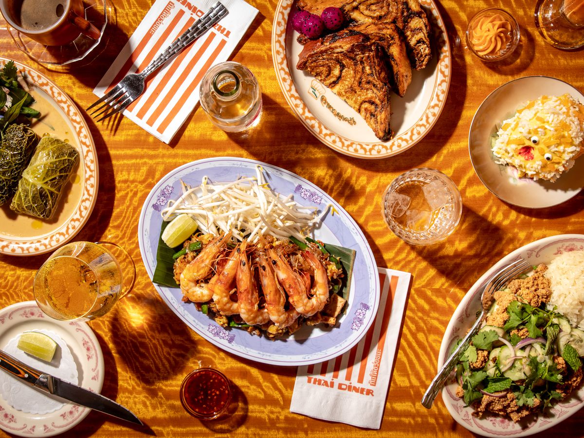 An aerial view of a spread of dishes, including vibrant cabbage rolls, pink head-on shrimp on phat Thai noodles, fried chicken larb, and a Thai tea pain perdu made with marbled babka.
