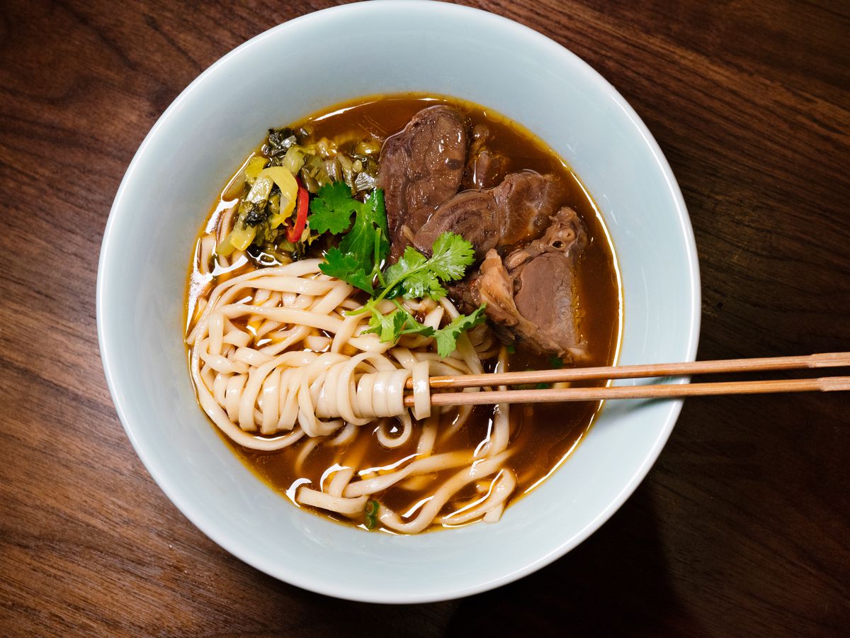 The image showcases a bowl of beef noodle soup from Ho Foods, with artfully arranged noodles surrounding chopsticks.